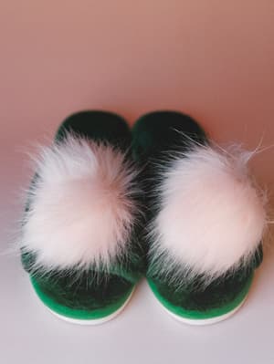 A pair of forest green fuzzy slippers with oversized blush pom-pom embellishments, shot from above against a warm dusty rose background — cozy and a little whimsical.