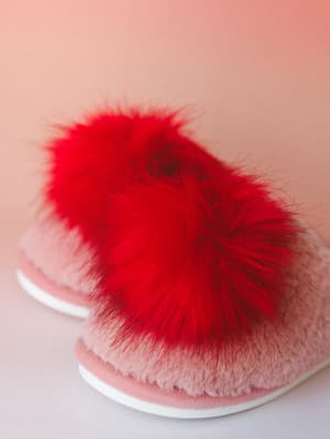 A pair of fluffy pink slippers with oversized red faux fur pom poms shot up close against a soft blush backdrop — the kind of thing that makes a Sunday morning feel like a whole event.