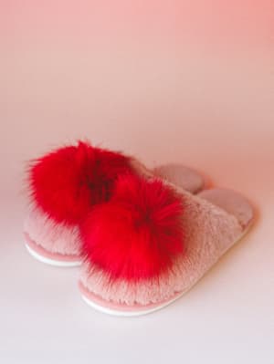 A pair of fluffy pink slippers with bold red pom-pom accents shot against a soft pink gradient backdrop — cozy and a little cheeky, like your favorite Saturday morning.