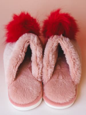 A pair of blush pink faux fur slippers shot from above against a soft pink background, topped with bold red pom pom trim — cozy and a little cheeky, like the best kind of Saturday morning.