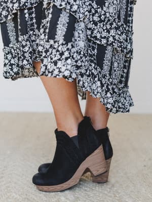 A close-up detail shot of black suede block-heel booties with a wood-stacked sole, styled with a flowy black and white floral midi skirt — the kind of pairing that makes a Tuesday feel intentional.