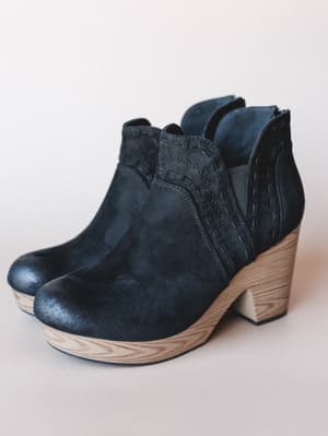 A pair of black suede platform ankle booties with a natural wood-grain heel and platform, detailed with woven stitching along the side panel — the kind of boot that makes every outfit feel a little more intentional.