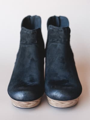 A pair of black suede platform booties shot straight-on against a soft neutral background — the wood-grain platform sole and tone-on-tone embossed ankle detail give them that lived-in, artisan feel.
