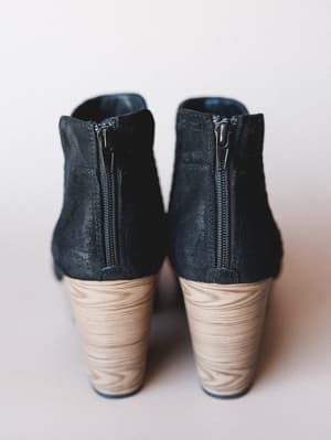 A close-up rear view of a pair of black suede ankle booties with wood-grain stacked heels and silver side zippers, shot against a soft neutral background — the kind of boot that does the heavy lifting on a Tuesday.