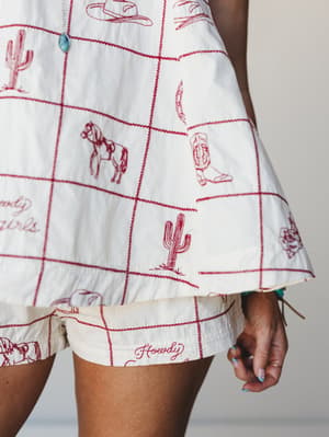 A close-up detail shot of a cream and red embroidered western-print matching set — a flowy top and shorts covered in grid lines and hand-drawn cowboy motifs like cacti, boots, horses, and 'Howdy Girls' script, finished with a turquoise button detail.