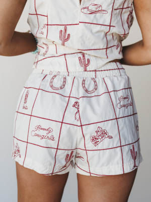 A close-up mid-shot of a cream matching set — cropped top and high-waist shorts — covered in red embroidered western motifs like cacti, horseshoes, cowboy hats, roses, and 'Howdy Cowgirls' script, framed by a red zig-zag grid stitch.