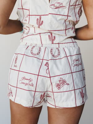 A close-up mid-shot of a cream matching set — cropped top and high-waist shorts — covered in red embroidered western motifs like cacti, horseshoes, cowboy hats, roses, and 'Howdy Cowgirls' script, framed by a red zig-zag grid stitch.
