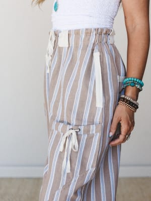 A close-up detail shot of striped wide-leg pants in a warm sandy tan with periwinkle blue and cream stripes, styled with a white textured top and layers of turquoise and beaded bracelets — relaxed and well-accessorized.