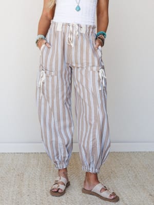 Striped harem-style pants in a warm tan and blue sit easy on the body — paired with a white crochet top, turquoise jewelry, and buckle sandals, the whole look feels like somewhere coastal and unhurried.