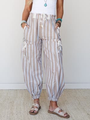 Striped harem-style pants in a warm tan and blue sit easy on the body — paired with a white crochet top, turquoise jewelry, and buckle sandals, the whole look feels like somewhere coastal and unhurried.