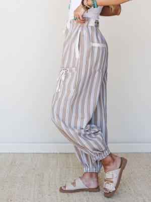 A side-profile shot of relaxed striped jogger pants in warm sand tones with periwinkle blue stripes, styled with a white textured top, turquoise stacked bracelets, and buckle sandals — easy, expressive, and ready for wherever the day goes.