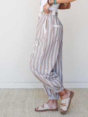 A side-profile shot of relaxed striped jogger pants in warm sand tones with periwinkle blue stripes, styled with a white textured top, turquoise stacked bracelets, and buckle sandals — easy, expressive, and ready for wherever the day goes.