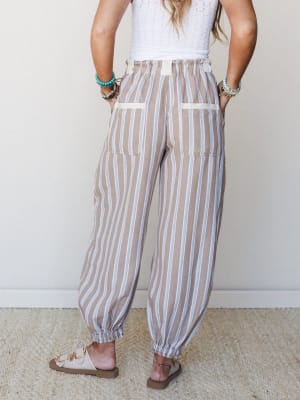 A back-facing model wears relaxed striped jogger-style pants in a warm taupe with white and blue pinstripes, paired with a white crochet top and neutral sandals — the kind of easy, put-together outfit that travels well and feels even better.