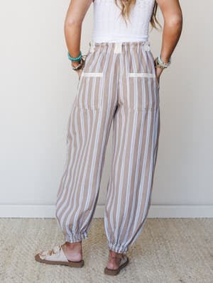 A back-facing model wears relaxed striped jogger-style pants in a warm taupe with white and blue pinstripes, paired with a white crochet top and neutral sandals — the kind of easy, put-together outfit that travels well and feels even better.