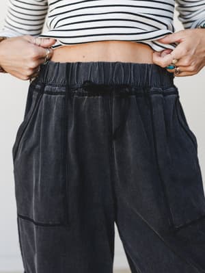 A close-up detail shot of wide-leg ash black pants with a washed, lived-in finish, styled with a cropped stripe top and layered rings — effortless and a little undone in the best way.