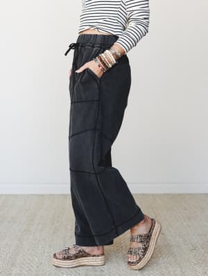 A side-profile shot of wide-leg ash black pants with visible seam paneling and a drawstring elastic waist, styled with a striped crop top, stacked bracelets, and leopard print platform sandals — the kind of outfit that looks effortless but clearly wa