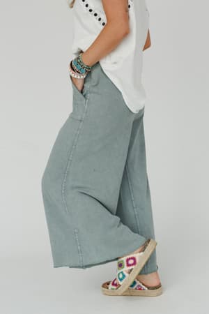 A person stands sideways wearing the Relaxing Robin Wide Leg Pant in Faded Teal, a white short-sleeve top, colorful beaded bracelets, and espadrille sandals with a multicolored crochet pattern.