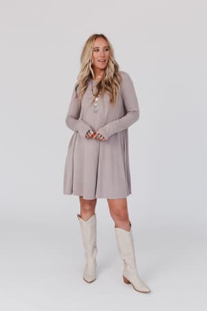 A woman with long blonde hair wears The Nest Jake Long Sleeve Crewneck Dress in Mocha Latte, styled with layered necklaces and cream cowboy boots. She stands against a white background, smiling slightly and gazing to the side.