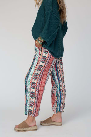 A person stands sideways with hands in pockets, wearing The Nest Staycation Wide Leg Drawstring Pant in Red Clay, a teal knit sweater, and tan espadrille sandals against a plain light background.
