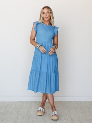 A cornflower blue tiered midi dress with smocked bodice and ruffle cap sleeves, styled with white platform espadrilles and stacked beaded bracelets — easy, warm-weather energy that works as hard as she does.