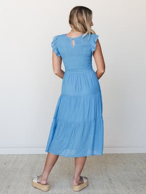 A breezy cornflower blue midi dress shot from the back, showing off a smocked bodice, flutter sleeves, and a tiered skirt that moves like it was made for a slow summer afternoon.