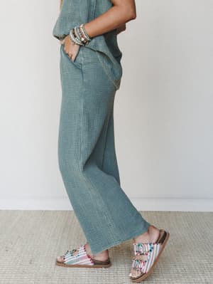 A side-profile shot focused on wide-leg gauze pants in a muted ash jade paired with matching top, colorful beaded bracelets, and boho fringe sandals — easy, expressive dressing that feels like a permanent vacation.