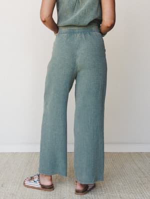 A back-view shot of relaxed wide-leg pants in a muted sage-teal gauze fabric, paired with a matching top and embellished sandals — the kind of easy set that feels like a vacation the moment you put it on.
