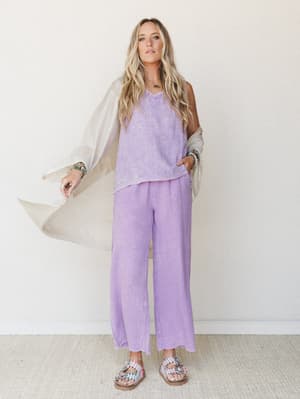 A breezy lavender matching set — gauze cami and wide-leg pants — styled with a draped oatmeal cardigan, stacked bracelets, and embellished sandals against a clean white studio wall; the whole look feels like a really good Saturday with nowhere to be.