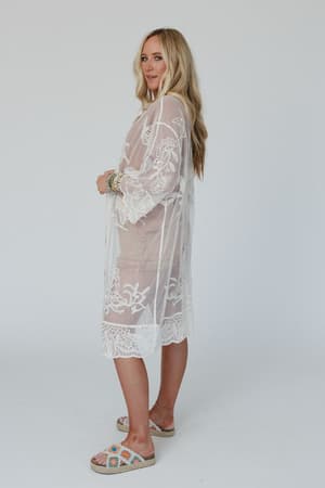 A woman with long blonde hair stands sideways against a plain light background wearing the Butterfly Beach Kimono - Ivory, a sheer white embroidered lace boho piece over a beige outfit with colorful slip-on sandals.