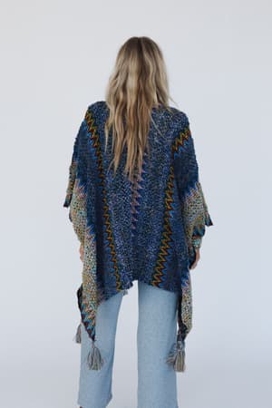 A person with long, wavy blonde hair wears the Desert Dreams Crochet Ruana in Cobalt with light-wash jeans—a perfect boho closet addition—while standing against a plain white background.