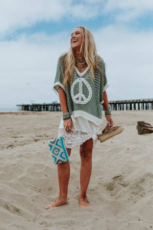 A barefoot woman with long blonde hair stands on a sandy beach, smiling. She wears the Prima Lace Hem Slip in Ivory under a green mesh top, holding sandals and a beaded clutch. A pier appears in the background.