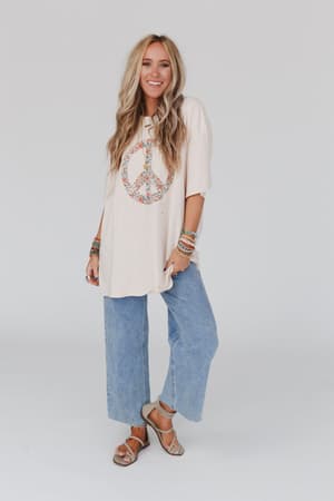 A woman with long, wavy blonde hair smiles in front of a white background, wearing the Jude Oversized Peace Sign Graphic Tee in oatmeal, light blue wide-leg jeans, tan sandals, and colorful bracelets.
