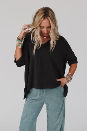 A woman with long blonde hair wears The Nest On The Go V Neck Ribbed Essential Tee in charcoal with light blue pants. She stands against a plain background, accessorized with colorful beaded bracelets, one hand in her pocket and the other touching her hair.