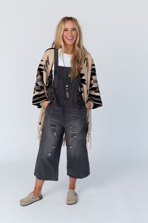 A woman with long blonde hair smiles while wearing the High Hopes Slouchy Pocket Denim Overalls with black pockets, a white shirt, beige and black patterned shawl, layered necklaces, and beige slip-on shoes against a plain white background.