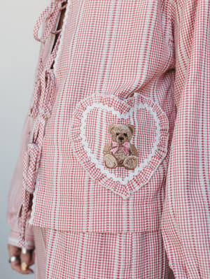 A close-up detail shot of a pink gingham two-piece set, showing a ruffled heart-shaped pocket with a sweet embroidered teddy bear — the kind of piece that feels like a hug you'd actually wear.