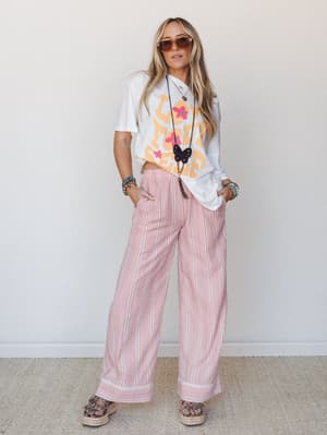 A relaxed full-body shot of a model in wide-leg gingham pants with stripe border trim, paired with an oversized graphic tee and layered boho accessories — the kind of outfit that looks effortless but is doing a lot of quiet work.