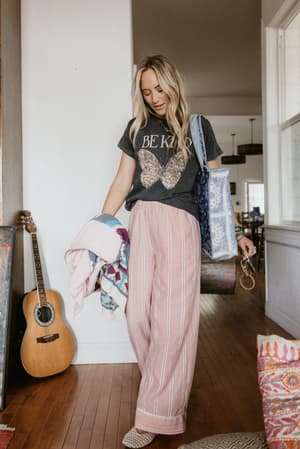 A relaxed, lived-in look shot inside a cozy home — graphic tee tucked into wide-leg striped pink pants, carried off with a patchwork tote and woven slides like she's heading somewhere unhurried.