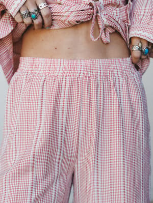 A close-up detail shot of a gingham wide-leg pant with a smocked waistband, styled with a matching tie-front crop top and stacked silver and turquoise rings — sweet, relaxed, and very much the kind of set you'd wear all day on purpose.