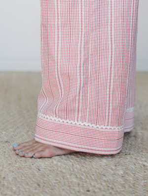 A close-up of wide-leg gingham pants with delicate ric-rac trim at the hem, shot barefoot on a jute rug — sweet, relaxed, and just a little nostalgic.