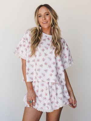 A matching floral two-piece short set in soft white with scattered pink rose clusters — styled with silver rings and pearl drop earrings, giving off an easy, feminine energy that lives somewhere between a lazy Sunday morning and a brunch you actually