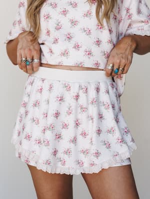 A close-up mid-shot of a matching floral two-piece set — a cropped tee and mini skirt with eyelet ruffle hem — styled with stacked silver and turquoise rings for that effortlessly personal touch.