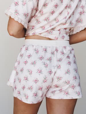 A back-facing close-up of a matching floral PJ set — waffle-textured shorts and a cropped top in cream with scattered pink rose clusters, finished with lace trim sleeves and an elastic waistband that makes the whole thing feel like a soft Saturday mo