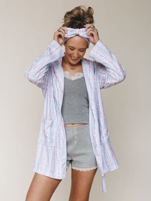 A smiling model adjusts a matching bow headband while wearing a soft floral-stripe robe over a gray lace-trim cami and button-front shorts — the whole look feels like a really good Saturday morning.
