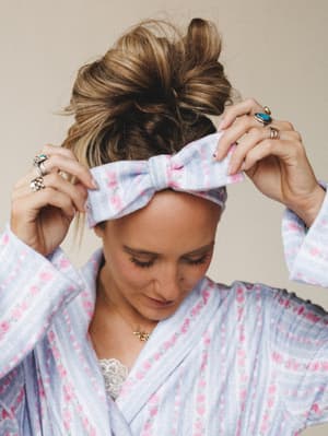 A soft, dreamy close-up of a woman adjusting a floral bow headband to match her robe — the kind of morning-routine moment that somehow looks effortlessly put-together.