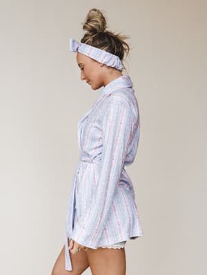 A model shot in profile wearing a floral-striped sleep robe in soft periwinkle and pink, belted at the waist and paired with a matching scrunchie headband and peeking lace shorts — the kind of morning look that makes staying in feel intentional.