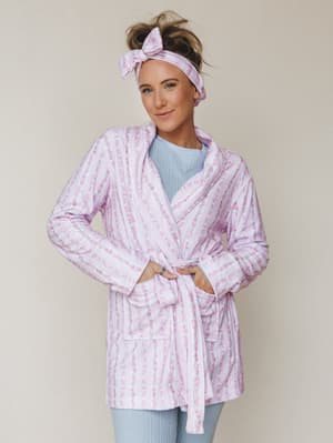 A smiling model wears a soft lavender-pink floral robe tied at the waist, layered over a light blue cable-knit top, finished with a matching bow headband — the kind of morning look that makes staying in feel intentional.
