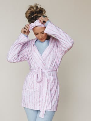 A model adjusts a matching floral headband while wearing a soft pink belted robe layered over a baby blue cable-knit top — the kind of morning look that makes staying in feel intentional.