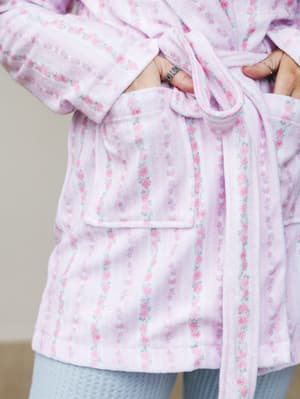 A close-up of a plush floral robe in the softest lavender-pink, hands tucked into the patch pocket with a tied sash at the waist — the kind of thing you wear on a slow Saturday morning and never want to take off.