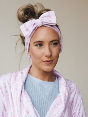 A close-up portrait of a model wearing a matching floral pink spa headband with a bow tied at the top, layered over a soft robe in the same print — the kind of morning routine moment that feels equally cozy and put-together.