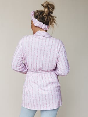 Shot from behind, a model wears a soft pink floral-stripe robe tied at the waist, paired with a matching scrunchie headband and light blue ribbed leggings — the kind of morning look that makes staying in feel intentional.